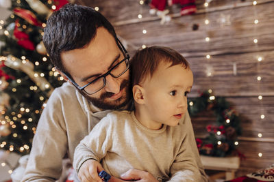 Daddy holding and playing with his son in living room decorated by christmas tree and present