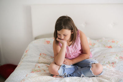 Girl child serious and frustrated sitting on a bed in a bright bedroom, concept age crisis 