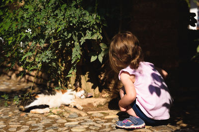Rear view of girl crouching by cat sleeping in yard