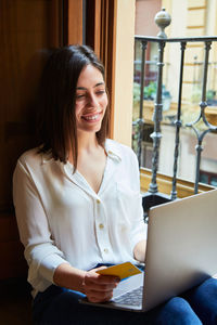 Young woman shopping and paying online on a laptop with credit card while relaxing at home next to a window.