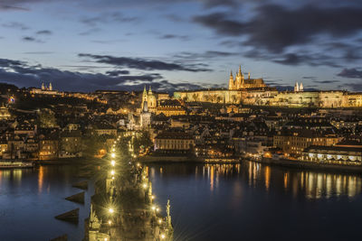 Illuminated buildings by river against cloudy sky