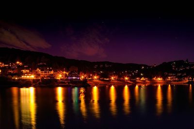 Illuminated city by river against sky at night