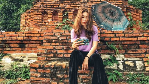 Young woman taking selfie with smart phone while sitting against brick wall