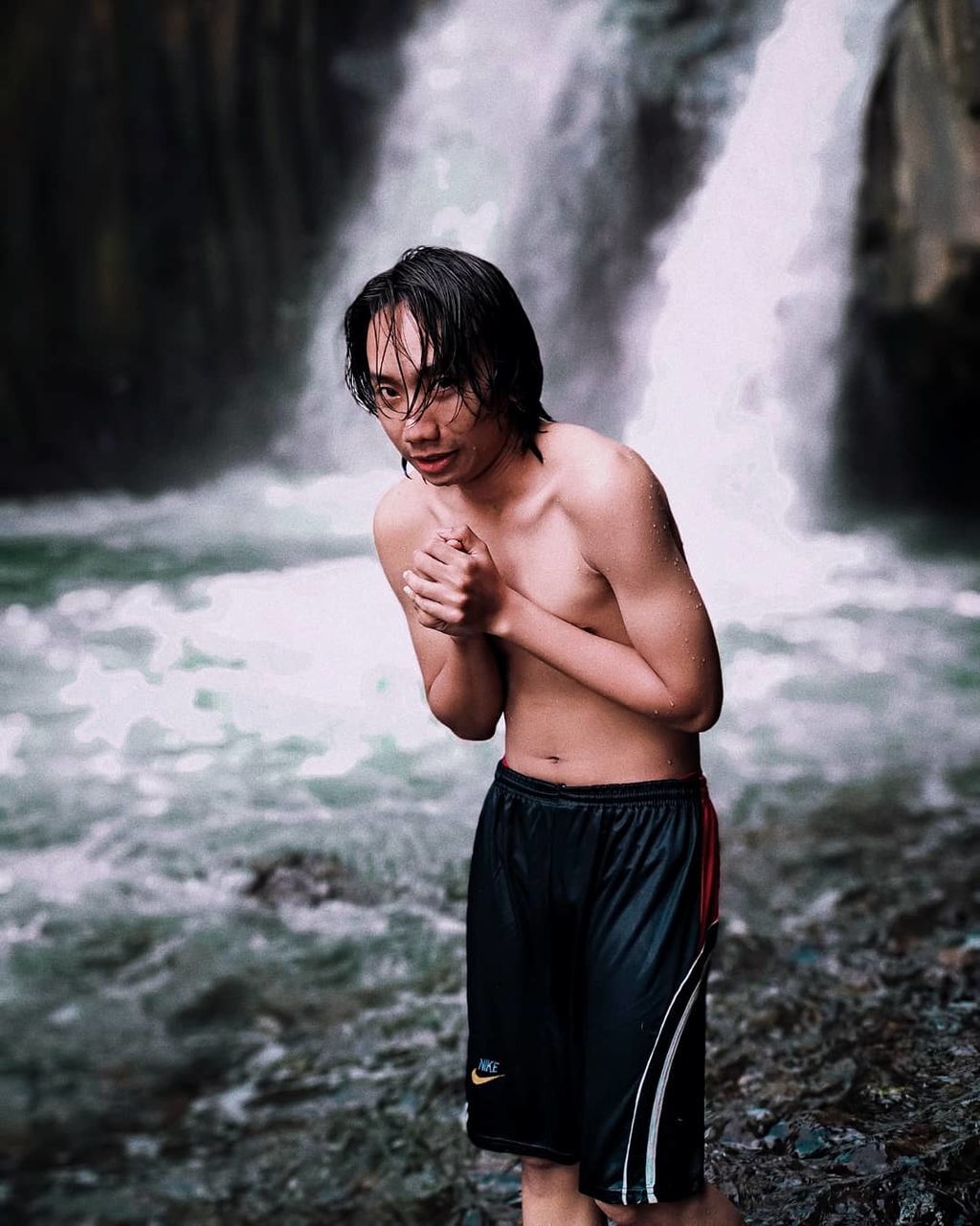 water, three quarter length, standing, real people, one person, lifestyles, motion, shirtless, leisure activity, young men, waterfall, focus on foreground, flowing water, young adult, day, nature, land, men, outdoors, shorts, power in nature