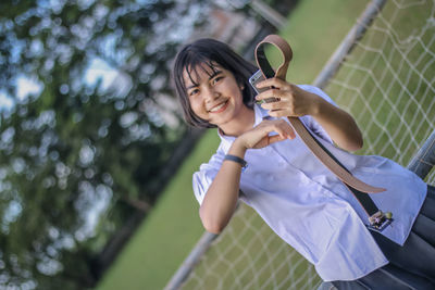 Portrait of smiling young woman holding smart phone and belt in park