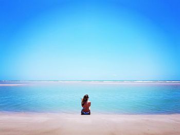 Rear view of woman looking at sea while sitting at beach against clear sky