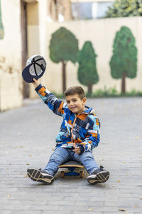 Portrait of smiling boy with cap sitting on skateboard at footpath