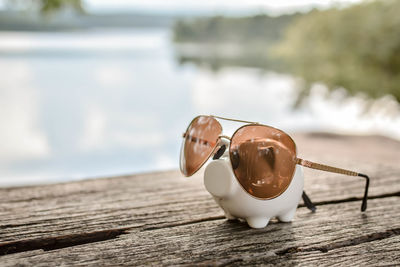 Close-up of sunglasses with piggy bank on wooden table against lake