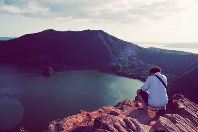 Rear view of man looking at lake while crouching on mountain against sky