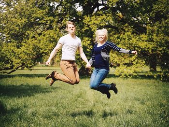 Cheerful young couple jumping against trees on grassy field