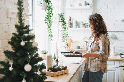 Attractive smiling woman with curly hair in plaid shirt bakes cookies at bright kitchen at home