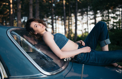 Hipster woman lying on vintage car in the forest.