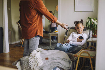 Midsection of father asking smart phone from daughter sitting on bed at home