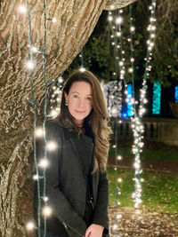 Portrait of smiling woman standing by illuminated tree