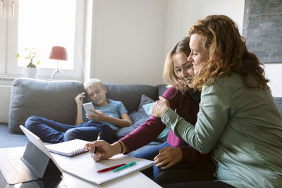 Mother embracing daughter studying while son using phone at home