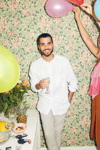 Portrait of smiling young man holding drink while standing by woman with balloons against wallpaper at home during dinne
