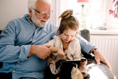 Girl using digital tablet while sitting with grandfather on sofa at home