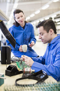 Male electrician soldering circuit board with colleague sitting on desk in industry