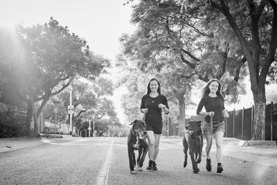 Portrait of smiling sisters with dogs running on road against trees