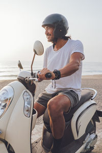 Side view of happy active guy dressed in white t shirt with shorts and black helmet riding scooter in summer evening on beach