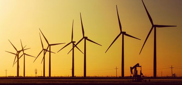 Oil well by wind turbines on field against sky during sunset