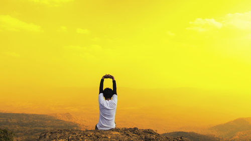 Rear view of man with arms raised sitting on mountain against orange sky