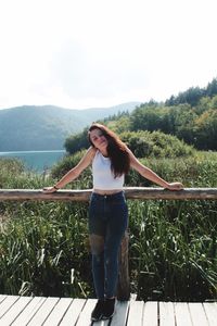 Portrait of young woman standing by railing against trees and sky