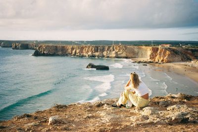 Rear view of woman sitting on rock by sea against sky