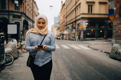 Young woman using smart phone while walking on street in city