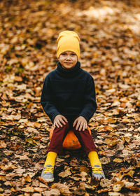 A boy a teen child in a bright yellow hat sits on a pumpkin in an autumn forest in nature outdoors