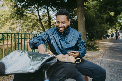 Happy male delivery person removing parcel from bag while sitting on bench at park