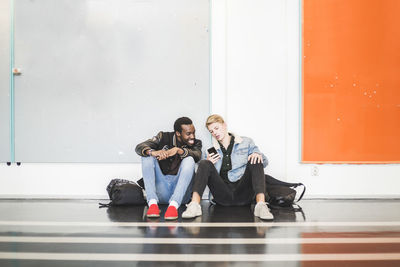 Young man showing mobile phone to friend while sitting in corridor of university