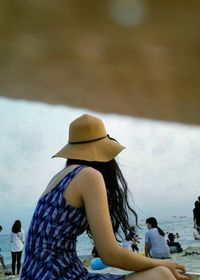 Side view of woman wearing hat sitting at beach against sky