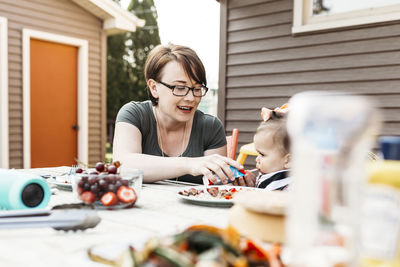 Mother feeding daughter while sitting at table in backyard