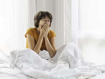 Sleepy woman is yawning in bed, covered with white blanket. it's hard to wake up early in morning.