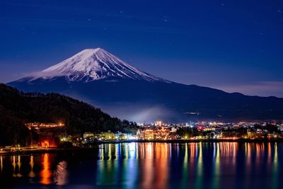 Illuminated city by river by snowcapped mountain at night