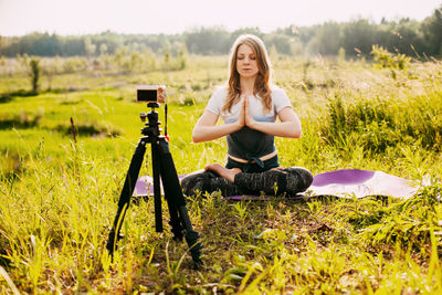 A young girl is doing yoga, sitting in the lotus position in nature and recording a video lesson 