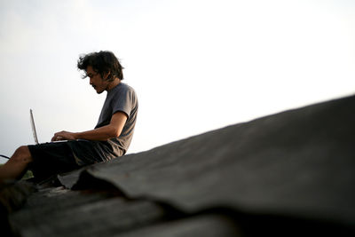 Side view of man using laptop against sky