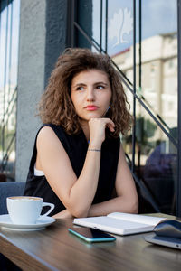 Young woman using notepad while sitting on table and drinking coffee