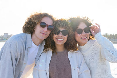 Group of young diverse women and man with curly hair and sunglasses embracing each other while standing on shoreline of cityscape in back lit