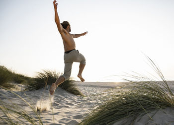 Shirtless young man jumping amidst plants at beach against clear sky during sunset