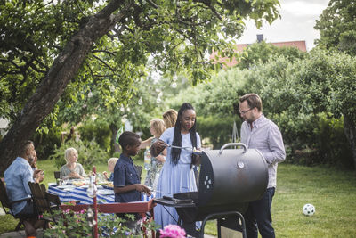 Man talking to woman with son preparing food at barbecue grill against friends during weekend party