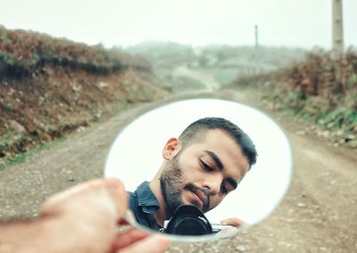 Cropped hand holding mirror with reflection of young man on road