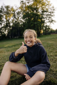 Portrait of happy girl showing thumbs up while sitting on grass in playground