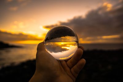 Close-up of hand holding crystal ball against sunset sky
