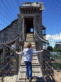 Rear view of boy walking on rope bridge against sky