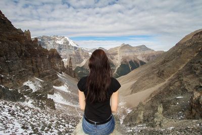 Rear view of woman sitting on cliff against mountains during winter