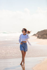 Full length of young woman standing at beach against sky