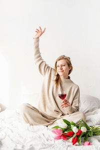 Woman sitting on the bed wearing pajamas, with pleasure enjoying flowers and a glass of red wine