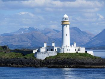 Lighthouse by sea and mountain against sky